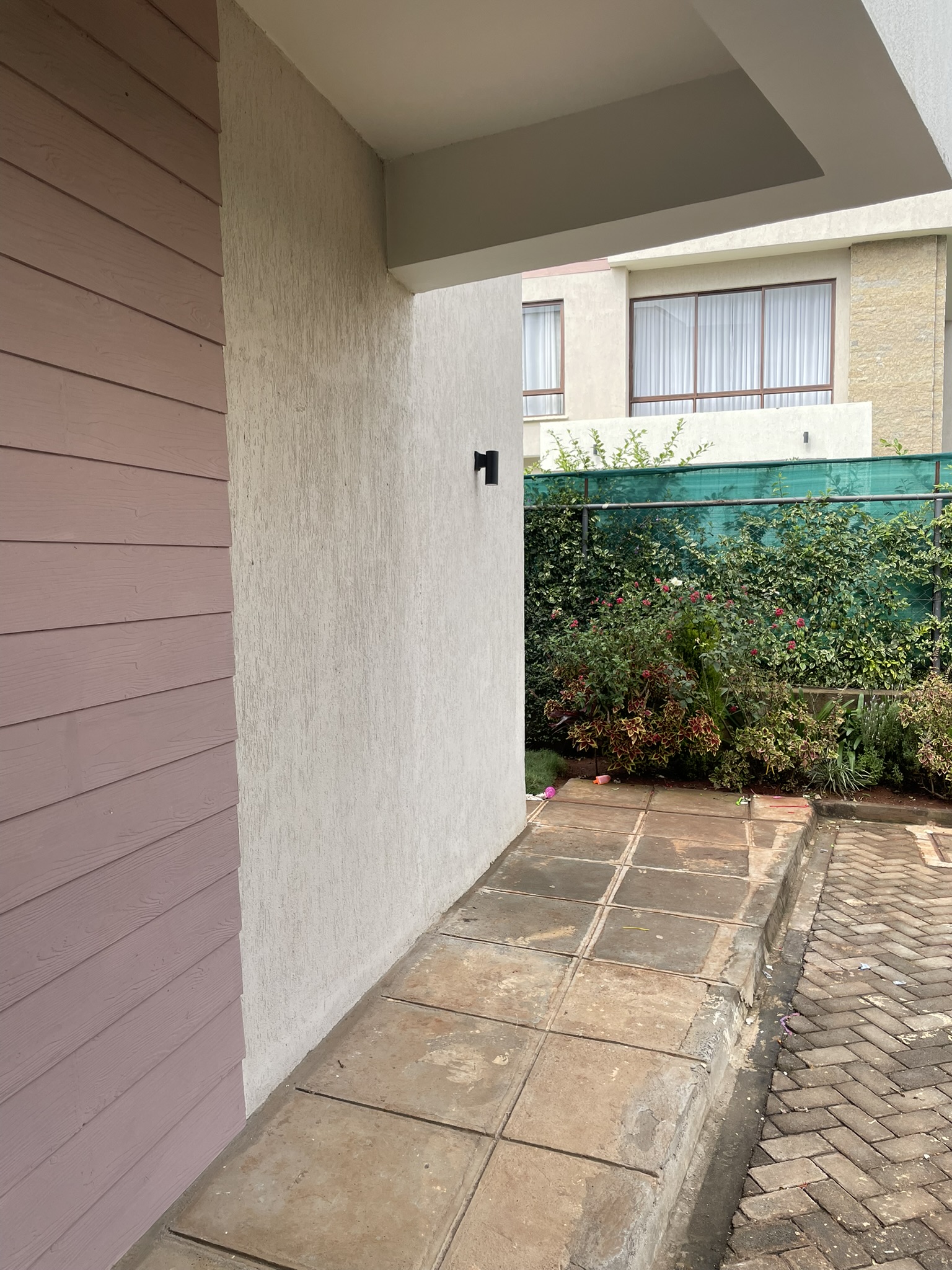 Outdoor paved walkway along the side of the house bordered by manicured hedge and black window grills.